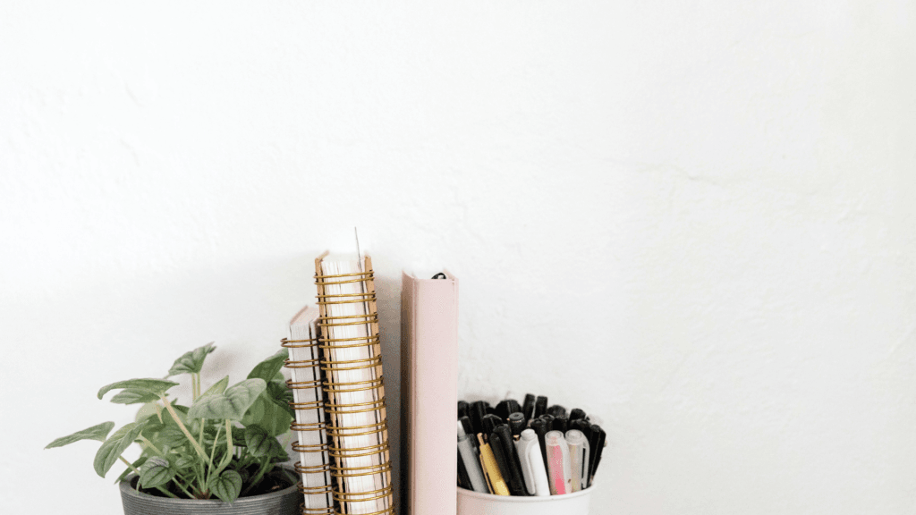 a copywriter desk, minimalist desk setup with a small potted plant, a stack of spiral notebooks with gold bindings, and a white cup filled with pens and markers, set against a plain white wall.